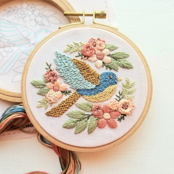 A round embroidery hoop holding a floral and bird design in pastel thread, photographed on a light surface with other embroidery threads and a pattern sheet partially visible.