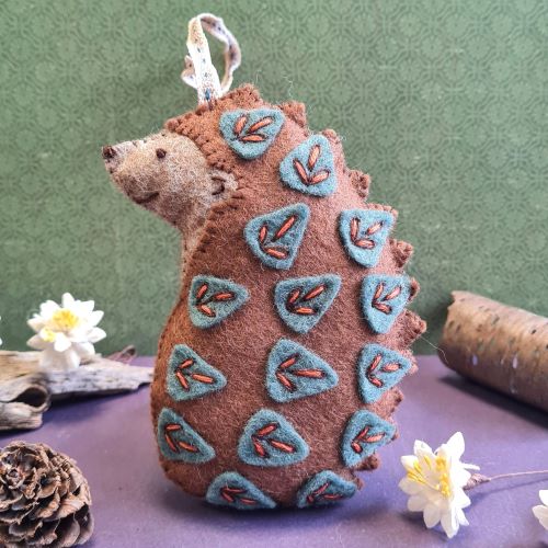 Hedgehog-shaped ornament with embroidered details on a purple surface with a green background. Designed by Corinne Lapierre. Available at tinypatio.com