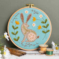 Felt Embroidery hoop with a rabbit design on a teal background, surrounded by flowers and spools of thread. By Corinne Lapierre. Available at tinypatio.com