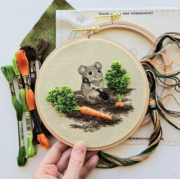 A hand holds a wooden embroidery hoop featuring a stitched mouse holding a small shovel, surrounded by embroidered green plants and a carrot on beige fabric. Embroidery floss skeins and a pattern sheet are arranged around the hoop on a white surface. Garden Mouse Embroidery Kit by Jessica Long | 6-inch (15cm)