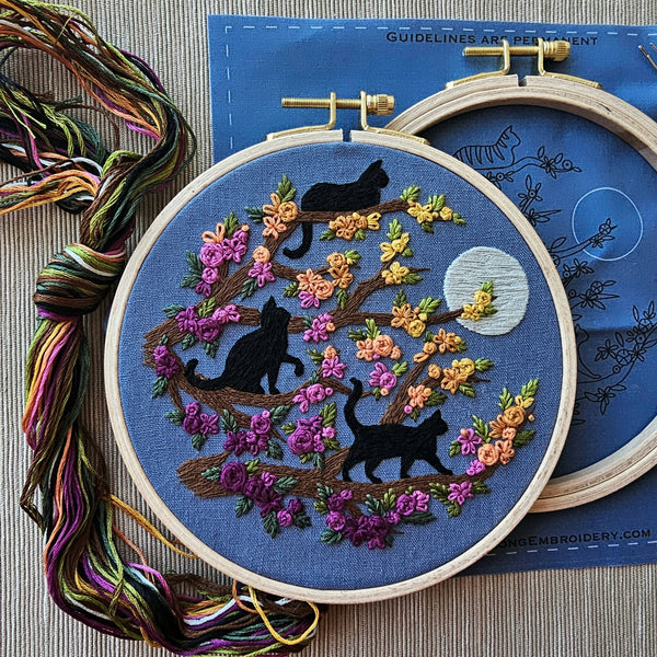 A modern embroidery hoop kit with black cat and floral design on blue fabric, surrounded by embroidery threads. By Jessica Long Embroidery