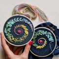 Two circular embroidery hoops with colorful floral and leaf stitched designs on dark fabric, held by a hand with pink nail polish against a light neutral background. Multicolored thread bundles and a blue fabric piece are visible.