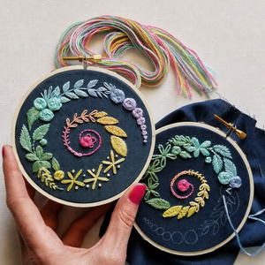 Two circular embroidery hoops with colorful floral and leaf stitched designs on dark fabric, held by a hand with pink nail polish against a light neutral background. Multicolored thread bundles and a blue fabric piece are visible.