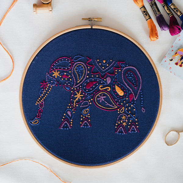 Finished paisley elephant embroidery in an 8 inch wooden hoop on white fabric, stitched in pink, purple and gold on navy
