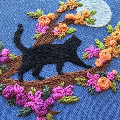 Embroidered design of a black cat among flowers on blue fabric. This is a Jessica Long embroidery kit designed for adults who love adorable cats.