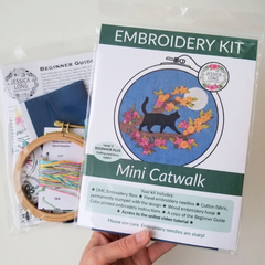 An embroidery kit package labeled 'Mini Catwalk' designed by Jessica Long Embroidery, held by a hand, with included materials partially visible behind it, including a wooden hoop with thread, a printed beginner guide, and a blue fabric piece on a white background.
