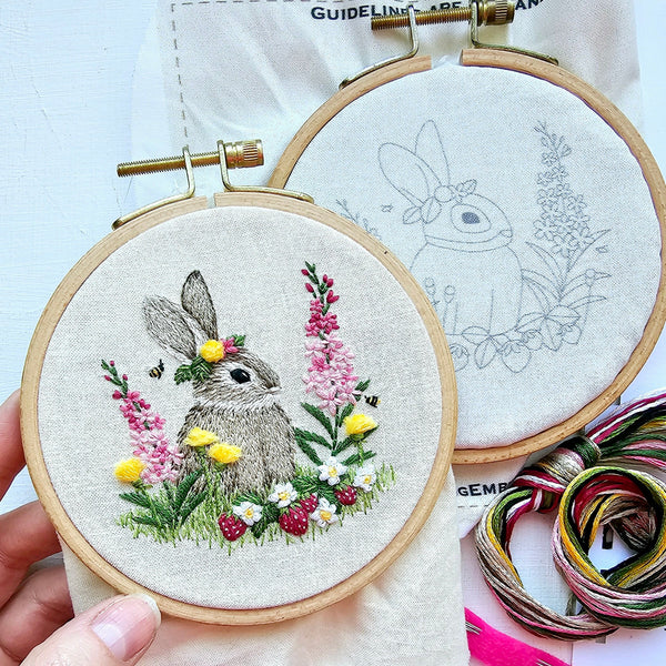 A close-up of an embroidery hoop holding a finished embroidered bunny scene with pink flowers, yellow flowers, green grass, and small bees on a white fabric background. A second hoop behind it shows a gray outline of the same bunny design, and thread spools are visible at the lower right.
