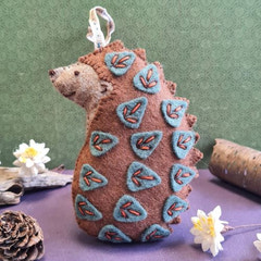 Hedgehog-shaped ornament with embroidered details on a purple surface with a green background. Designed by Corinne Lapierre. Available at tinypatio.com