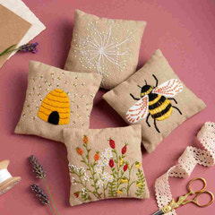 Four embroidered pillows with bee, flower, and honeycomb designs on a pink background.