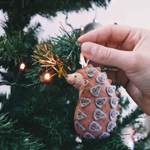 A hand is holding a felt hedgehog Christmas ornament decorated with blue and red leaf patterns, hanging from a green Christmas tree with warm string lights.