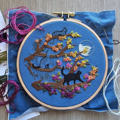 Embroidery hoop with an adorable cat-themed design on blue fabric, surrounded by embroidery threads. Designed by Jessica Long Embroidery. Curated by Tiny Patio in the UK. 