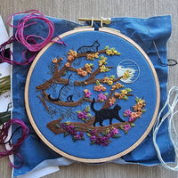 Embroidery hoop with an adorable cat-themed design on blue fabric, surrounded by embroidery threads. Designed by Jessica Long Embroidery. Curated by Tiny Patio in the UK. 