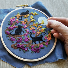 A hand holding an embroidery needle, stitching the catwalk beginner Embroidery kit. The design features three black cats and colourful flowers on blue fabric. Designed for adults by Jessica Long Embroidery.