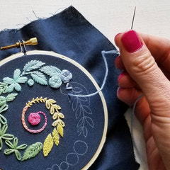 Jessica Long Spiral Sampler beginner embroidery hoop kit with floral design on blue fabric, hand holding a needle.