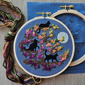 A modern embroidery hoop kit with black cat and floral design on blue fabric, surrounded by embroidery threads. By Jessica Long Embroidery