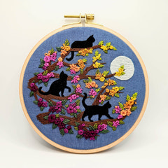 An Adorable Embroidery hoop kit with three black cats on branches, colorful flowers, and a moon on a blue background. Designed by Jessica Long Embroidery. Curated by Tiny Patio in the UK. 