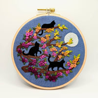 An Adorable Embroidery hoop kit with three black cats on branches, colorful flowers, and a moon on a blue background. Designed by Jessica Long Embroidery. Curated by Tiny Patio in the UK. 