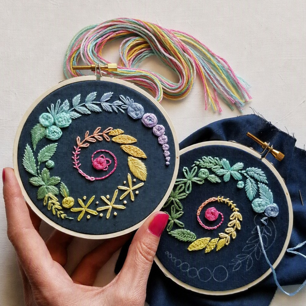Two circular embroidery hoops with colorful floral and leaf stitched designs on dark fabric, held by a hand with pink nail polish against a light neutral background. Multicolored thread bundles and a blue fabric piece are visible.
