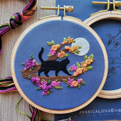 Close up of a 4-inch beginner embroidery hoop featuring a black cat sitting on a crescent moon, stitched on navy blue fabric. Modern Embroidery hoop with a black cat design on blue fabric, surrounded by colorful flowers and leaves. By Jessica Long Embroidery. 