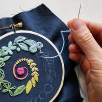 Jessica Long Spiral Sampler beginner embroidery hoop kit for adults with floral design on blue fabric, hand holding a needle.