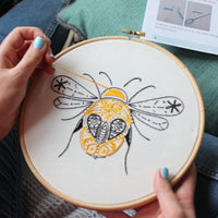 A hand stitching the bee's wing, drawing thread through the pale fabric in the hoop