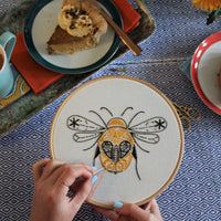 The bee embroidery in progress on a patterned cloth, with cake and a mug nearby