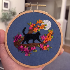 A hand holding a 4 inch wooden embroidery hoop with a finished black cat, colourful flowers, full moon on blue fabric, set against a blurred indoor background. Designed by Jessica Long Embroidery