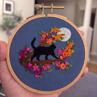 A hand holding a 4 inch wooden embroidery hoop with a finished black cat, colourful flowers, full moon on blue fabric, set against a blurred indoor background. Designed by Jessica Long Embroidery