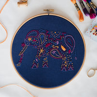 Finished paisley elephant embroidery in an 8 inch wooden hoop on white fabric, stitched in pink, purple and gold on navy