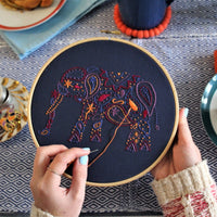 A pair of hands stitching the elephant design, pulling orange thread through the navy fabric in the hoop