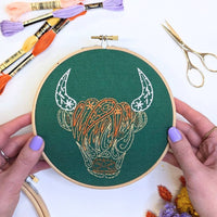 A pair of hands holding the finished Highland coo hoop above a white table with dried flowers and gold scissors