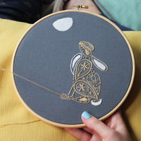 Hands holding the finished hare hoop against a yellow cloth, a length of gold thread still attached
