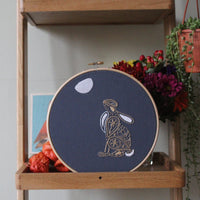 The hare embroidery displayed in a wooden shelf unit beside dried flowers