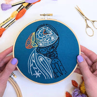 A pair of hands holding the finished puffin hoop above a white table with dried flowers and gold scissors