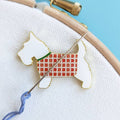 Enamel Scottish terrier needle minder in a red tartan coat, holding an embroidery needle and blue thread on hooped fabric