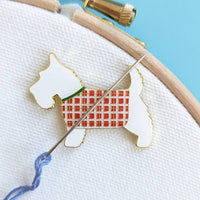 Enamel Scottish terrier needle minder in a red tartan coat, holding an embroidery needle and blue thread on hooped fabric