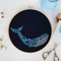 Finished blue whale embroidery in an 8 inch wooden hoop, laid on white linen with gold scissors and a reel of blue thread