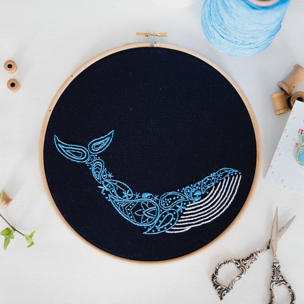 Finished blue whale embroidery in an 8 inch wooden hoop, laid on white linen with gold scissors and a reel of blue thread