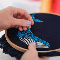 A hand pulling bright blue thread through navy fabric while stitching the whale's tail in the hoop