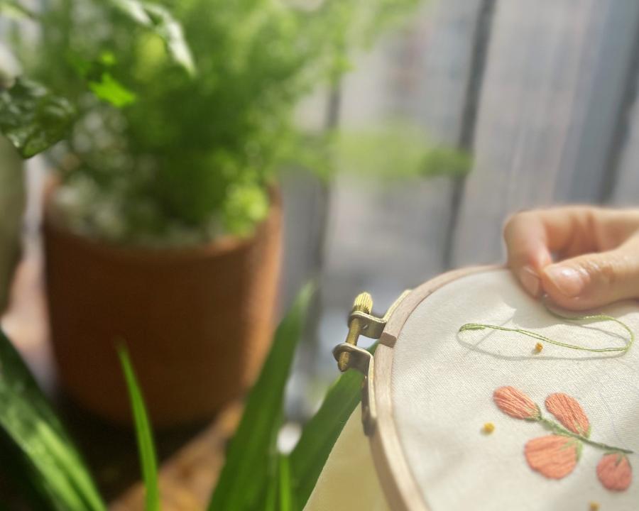 A hand stretching linen in an embroidery hoop beside a sunlit fern, with coral floral stitching in progress
